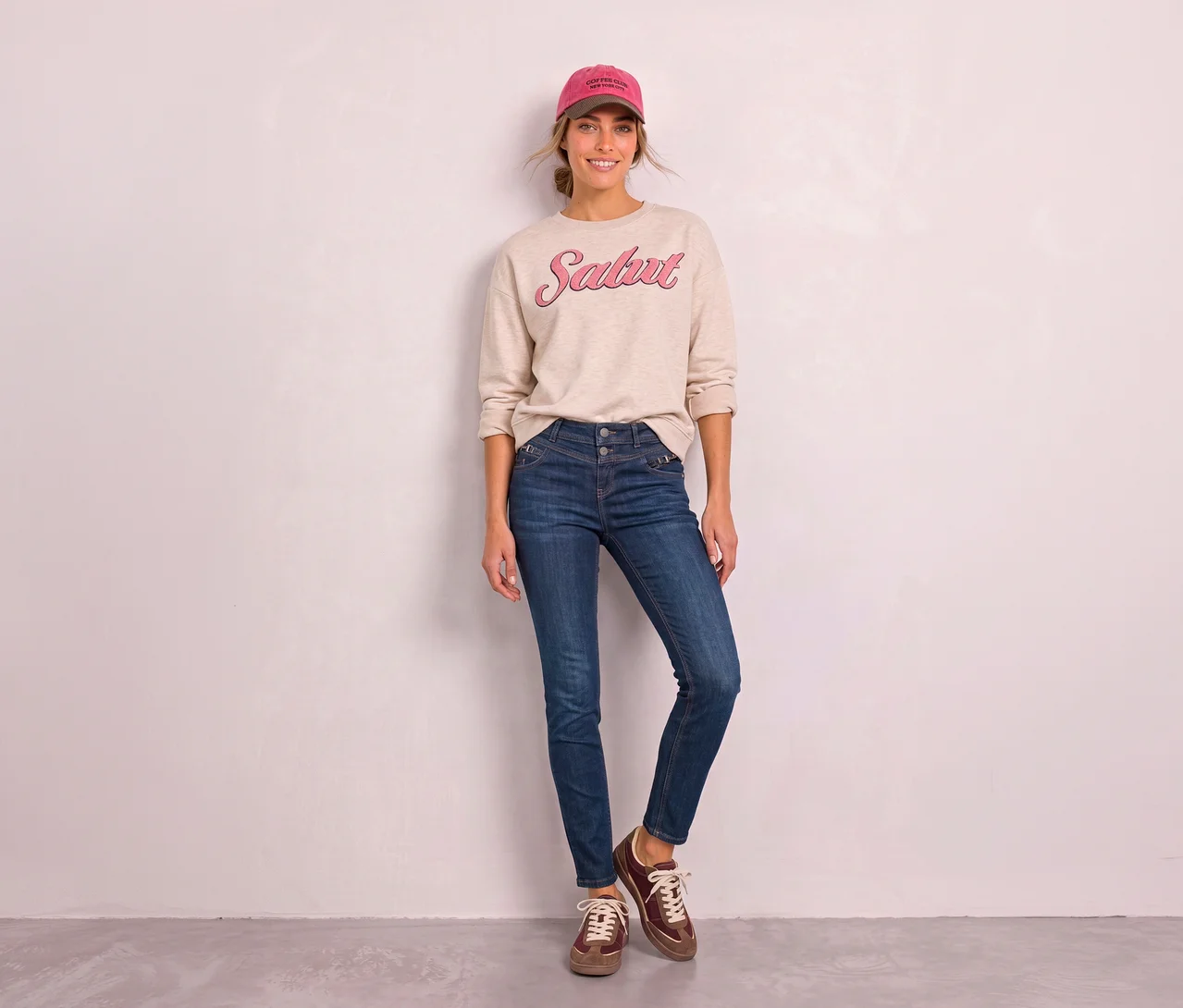 Eine lächelnde Frau mit rosa Baseballkappe, beigem Sweatshirt mit „Salut“-Aufschrift und blauen Street One Jeans steht vor einer hellen Wand.