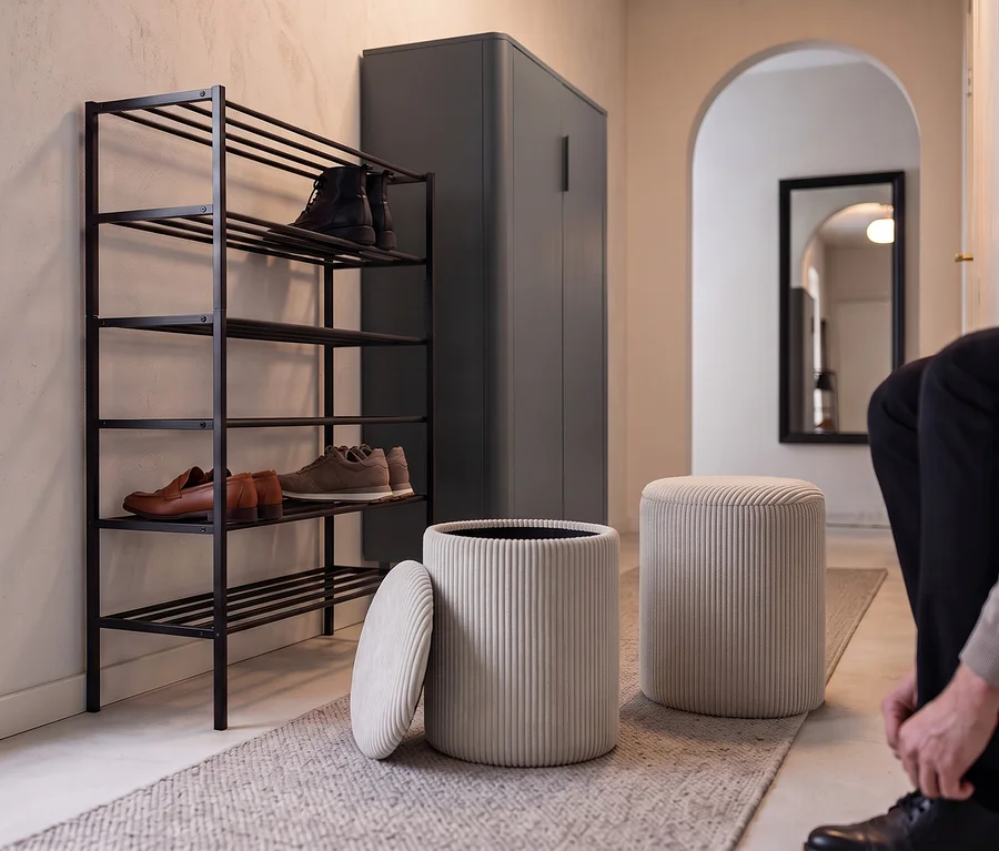 Ein Mann sitzt auf einem beigen Eglo Hocker »Vavatenina« und bindet sich den Schuh, neben einem WOOOD Schuhregal »Ferro« mit Schuhen und einem mattschwarzen WOOOD Schrank »Gunnar«.