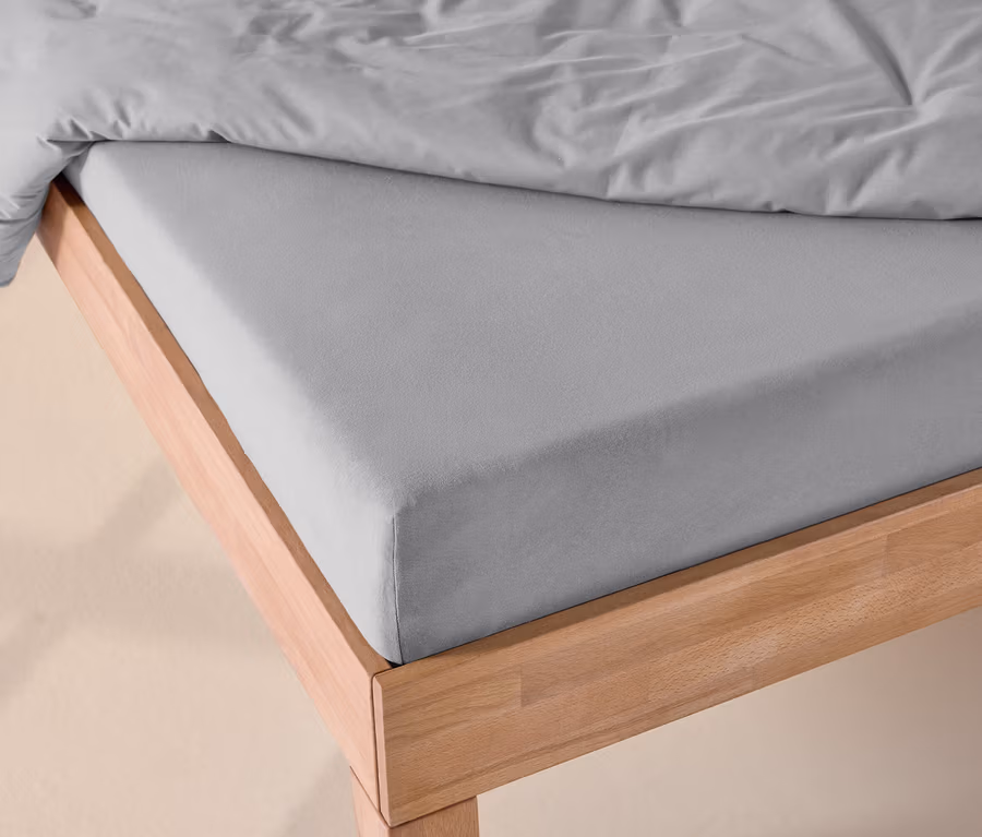Ein Bett mit einem grauen Jersey-Spannbettlaken auf der Matratze und einer grauen Bettdecke auf einem hellen Holzrahmen.