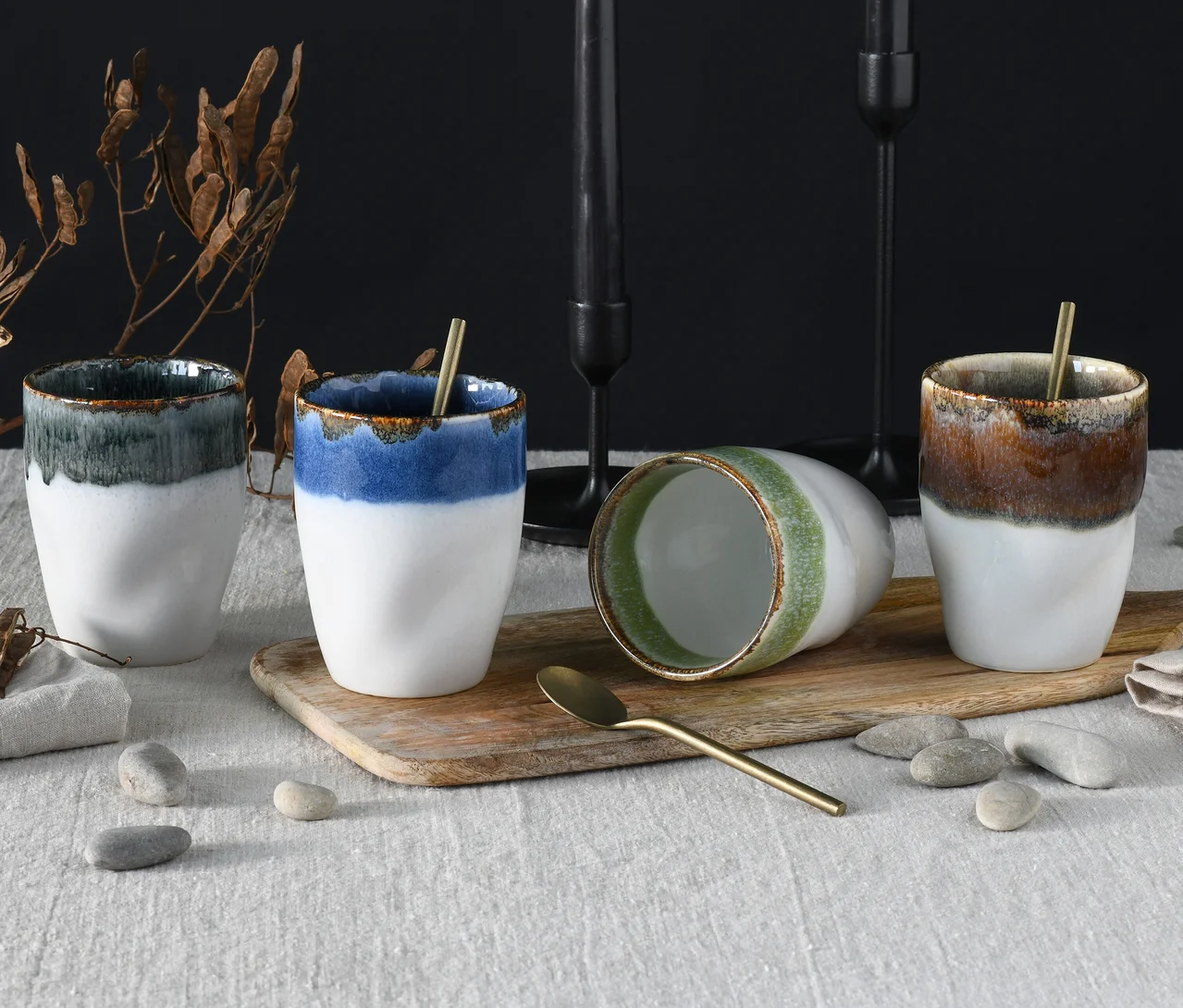 Vier creaTable Kaffeebecher aus dem Set „Squeezy Cacade“ stehen auf einem Holzbrett. Drei aufrechte Becher haben weiße Unterseiten und glasierte Oberseiten in Dunkelgrün, Blau und Braun. Ein grün glasierter Becher liegt auf der Seite. Zwei Becher enthalten goldene Löffel. Das Brett liegt auf einem weißen Tischtuch, umgeben von Kieselsteinen, getrockneten Blättern und schwarzen Kerzenhaltern im Hintergrund.