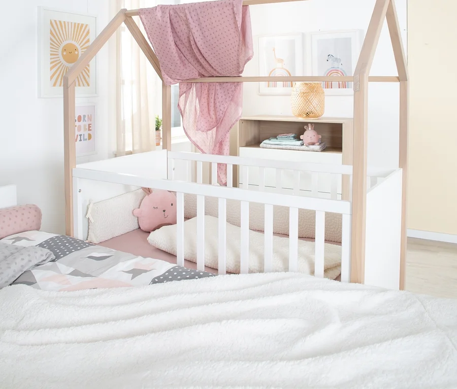 Kinderzimmer mit weißem und hölzernem Hausbett, über dem ein rosa Tuch hängt. Darin ein Gitterbett mit rosa Hasenkissen. Daneben ein Bett mit gemusterter Bettwäsche und weißer Flauschdecke.