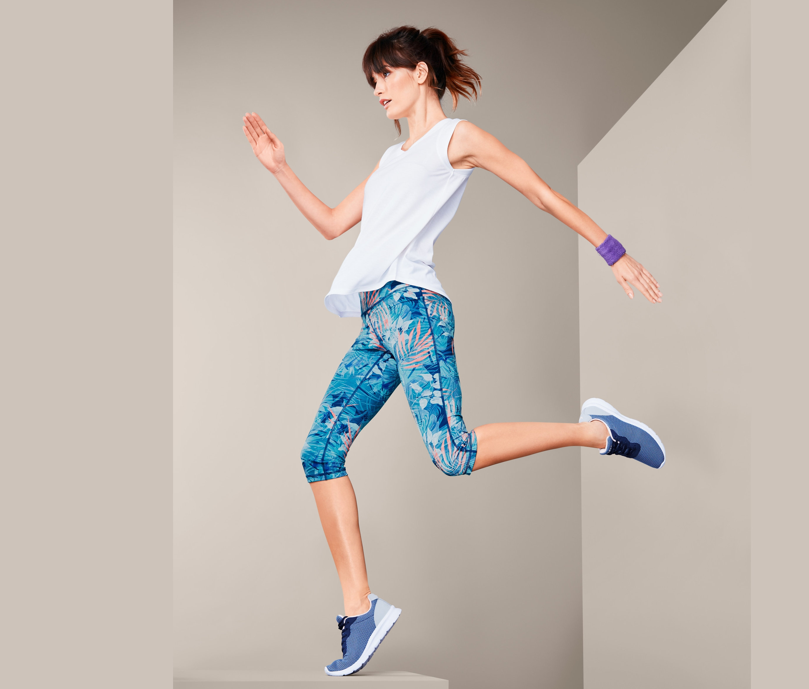 Frau springt und trägt eine 3/4-Sporttight in Dunkelblau, ein weißes Tanktop und blaue Turnschuhe.