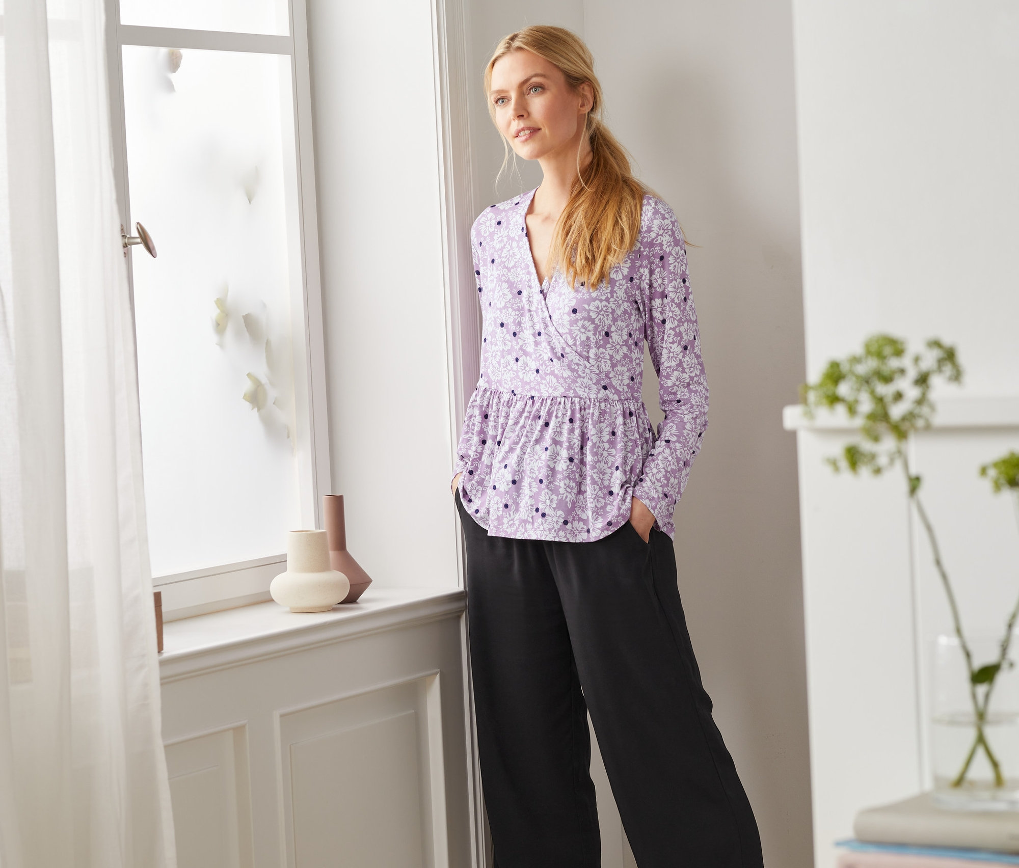 Frau steht am Fenster und trägt ein Langarmshirt.