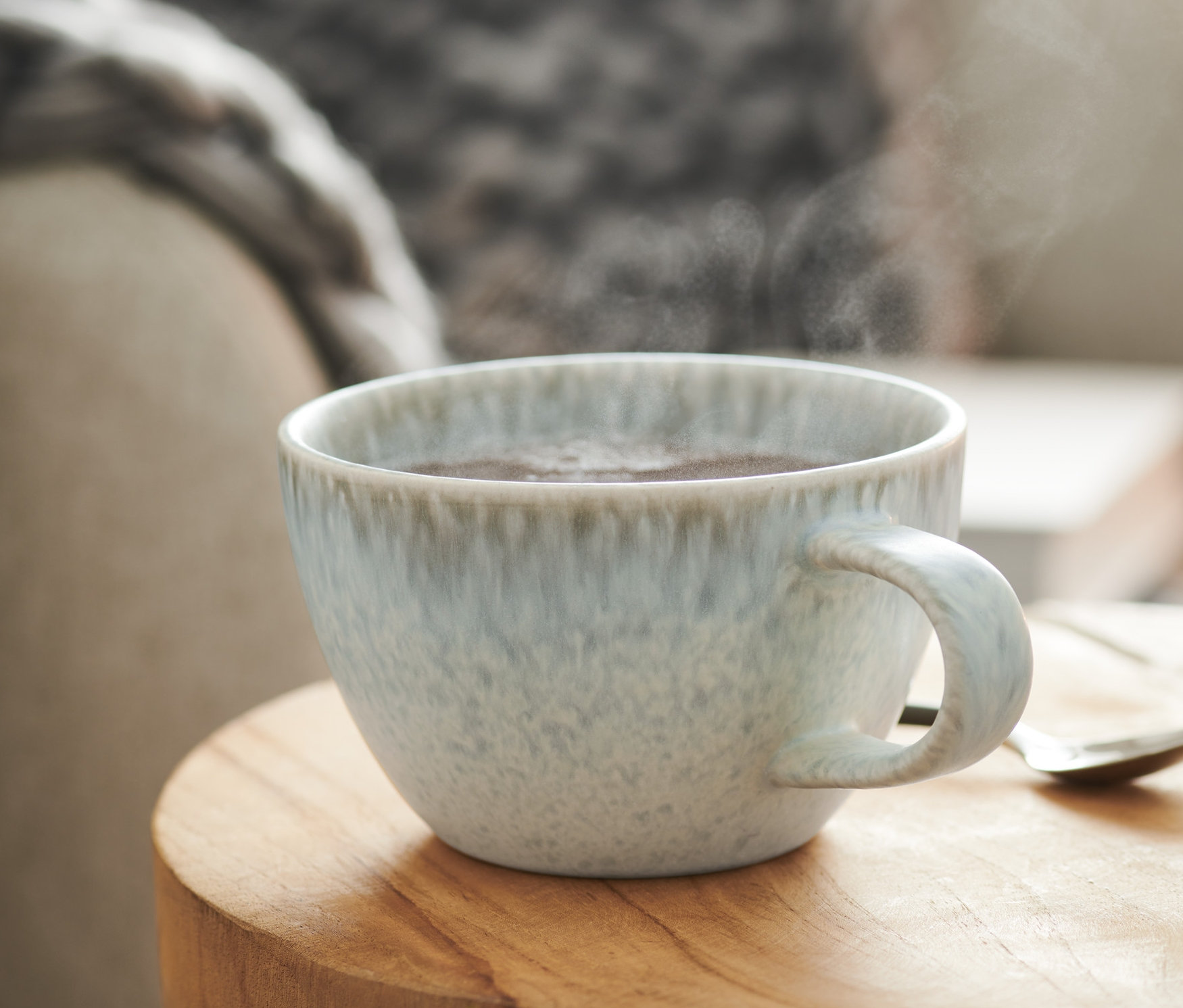 Dampf steigt aus der Jumbotasse auf einem Holztisch neben einem Hocker.