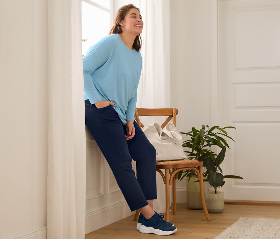 Eine Frau lehnt am Fenster und trägt Leder-Sneaker, dunkelblaue Joggpants, ein Fledermausshirt und einen Canvas-Shopper.