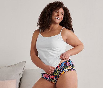 Frau kniet auf einem Bett und hält ein weißes Hemdchen und 5 Pantys.