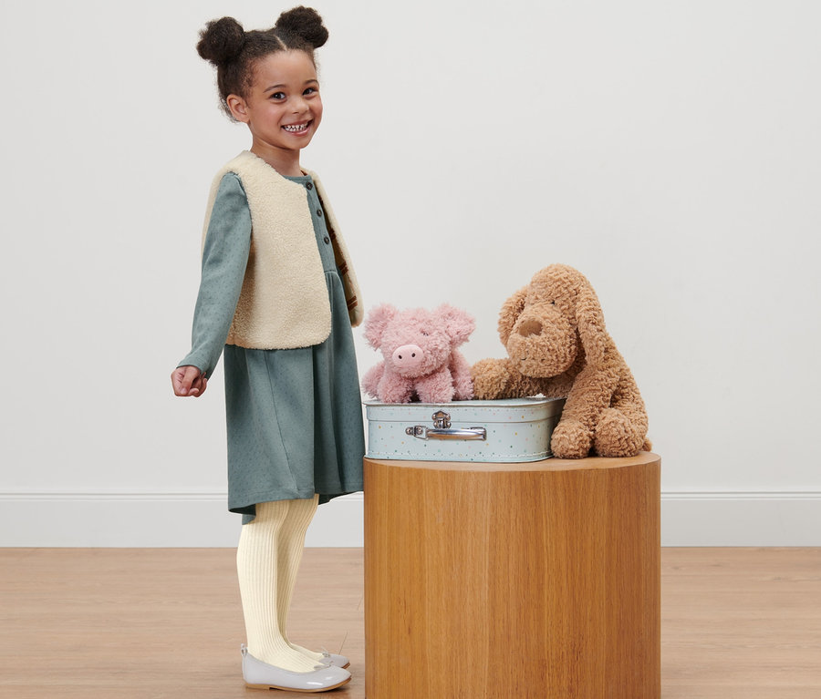 Lächelndes kleines Mädchen steht neben einer Kindertruhe mit Stoffschwein und Hund, trägt ein Kleinkind-Kleid und eine Kleinkind-Teddyweste.