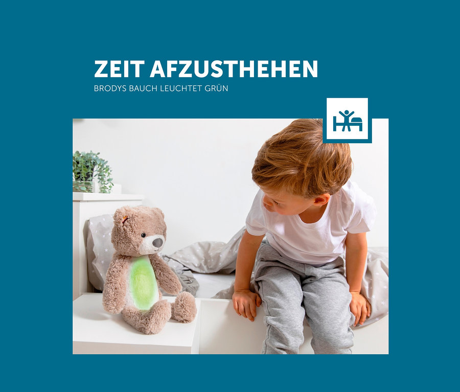 Ein Junge sitzt neben dem Zazu Nachtlicht »Brody der Bär« auf dem Bett.