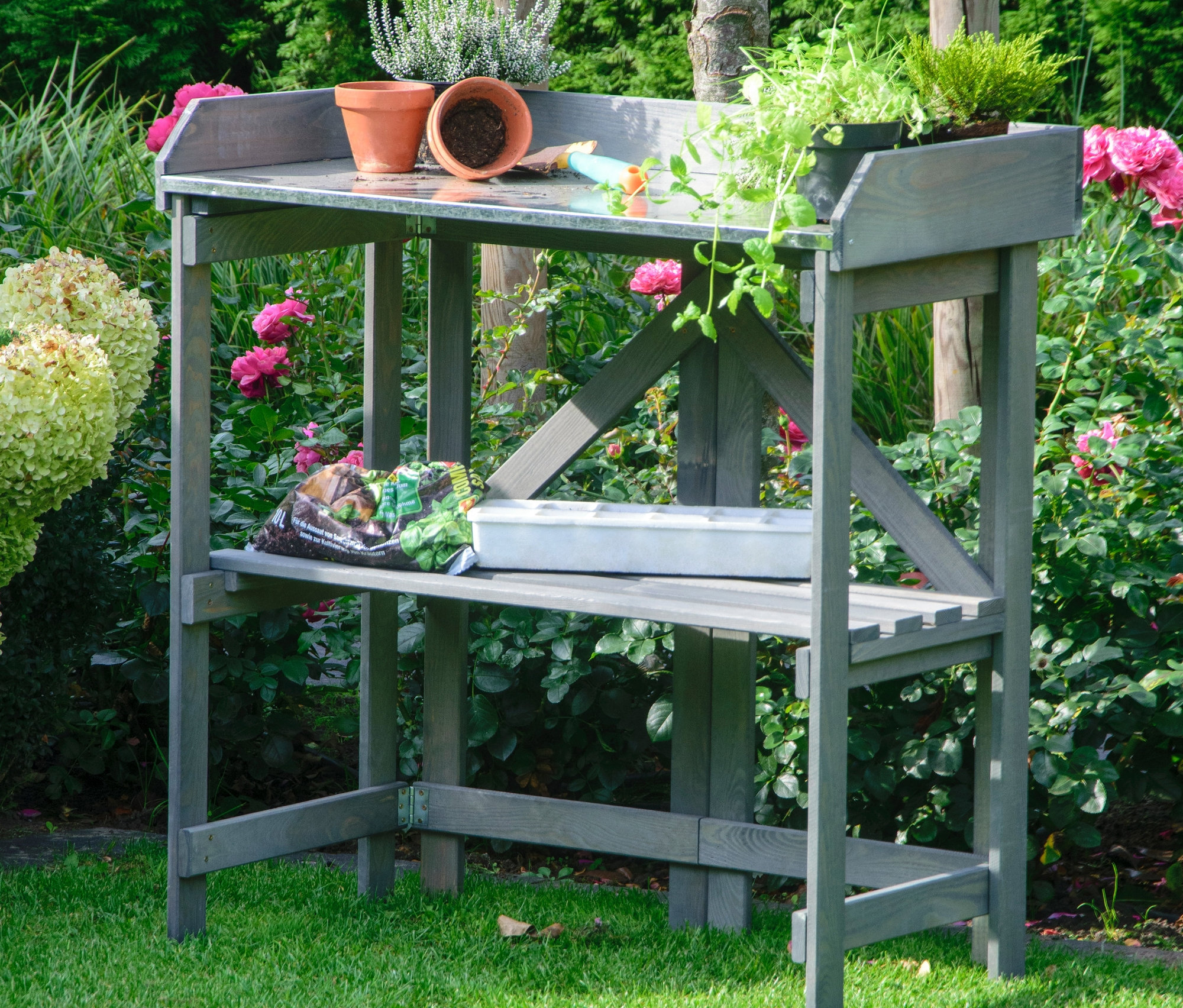 Ein grauer, klappbarer Pflanztisch mit Blumen und Töpfen auf der oberen Ablage und Anzuchtschalen auf der unteren.