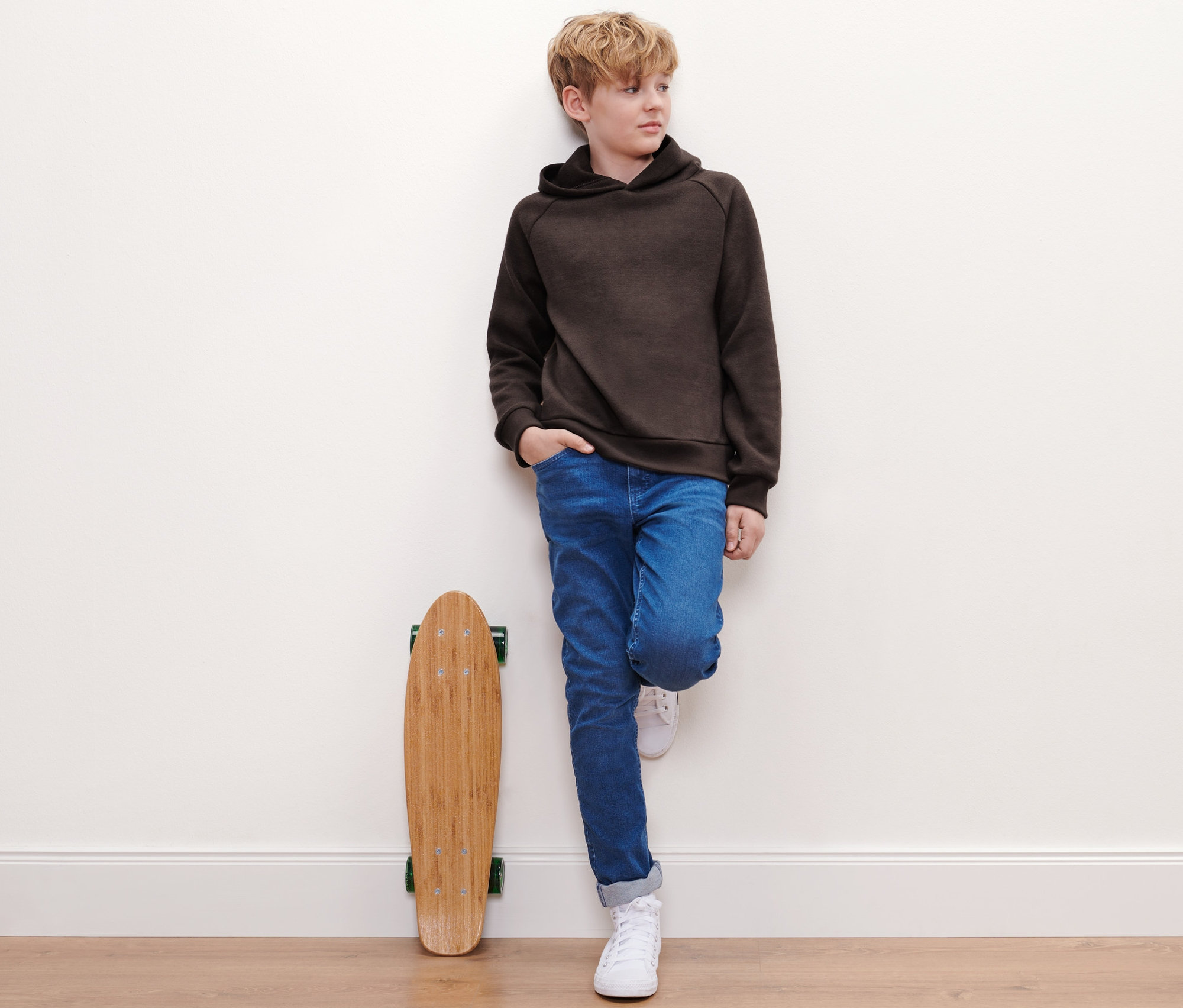 Junge lehnt an Wand, trägt Kinder-Strickhoodie, Jeans und Turnschuhe. Skateboard steht neben ihm.