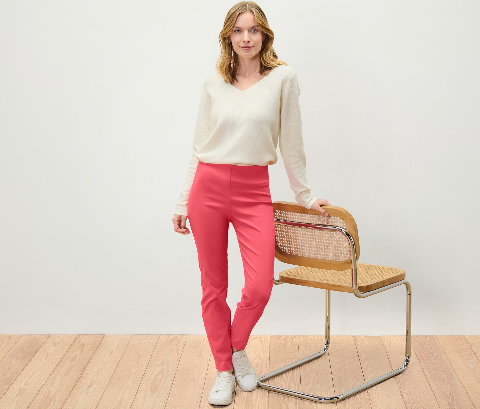 Frau steht an einem Stuhl angelehnt und trägt eine koralle Stretchhose.