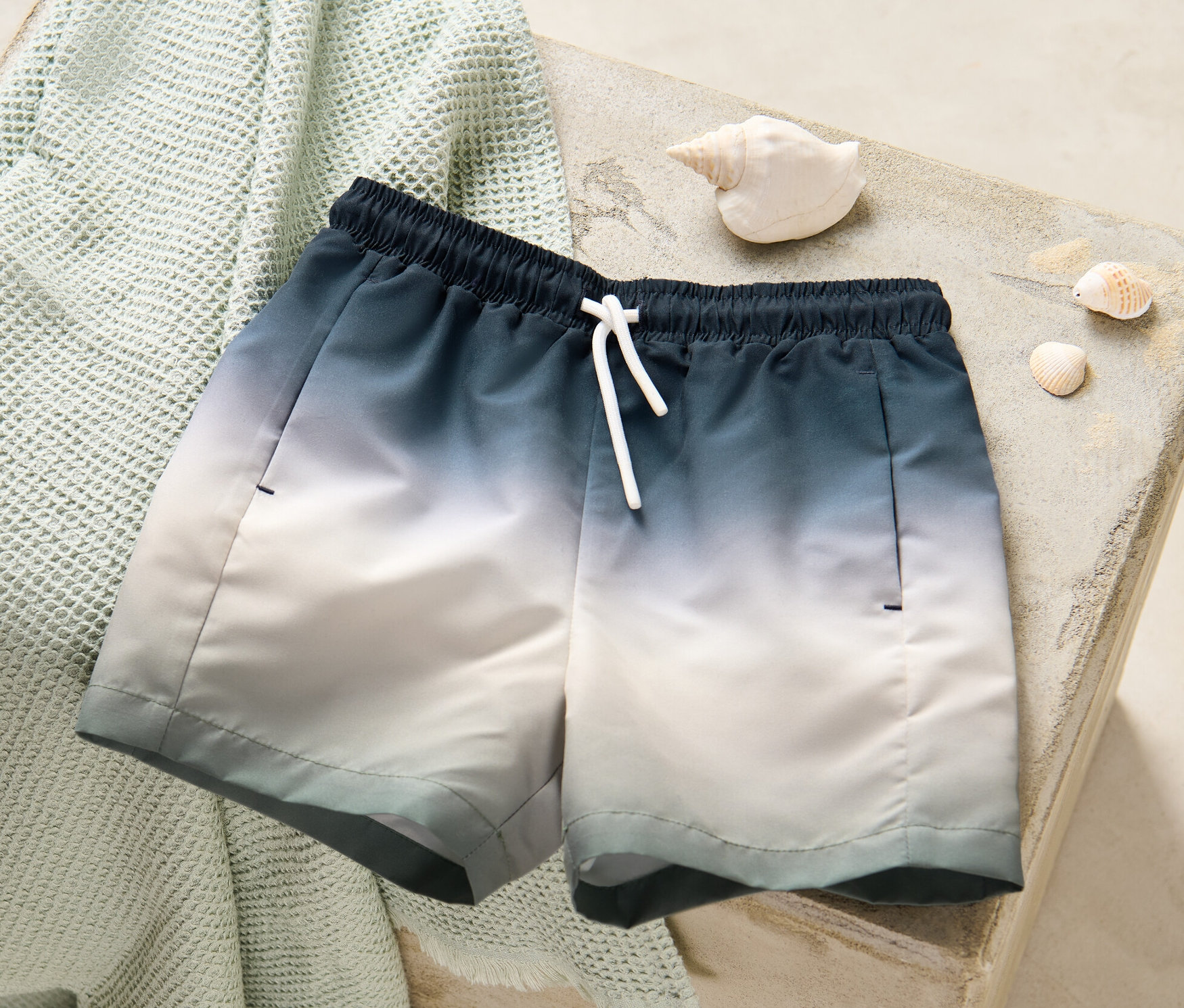 Kinder-Boardshorts liegen auf einem gefalteten Handtuch neben Muscheln.