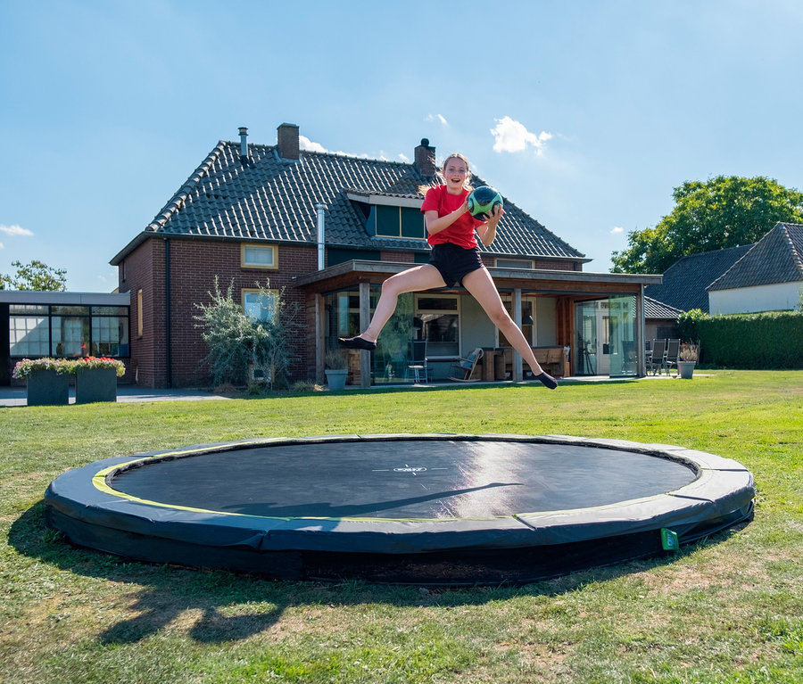 Eine junge Frau springt auf einem EXIT Bodentrampolin »Silhouette sports«, Ø ca. 366 cm und hält einen Ball im Garten.