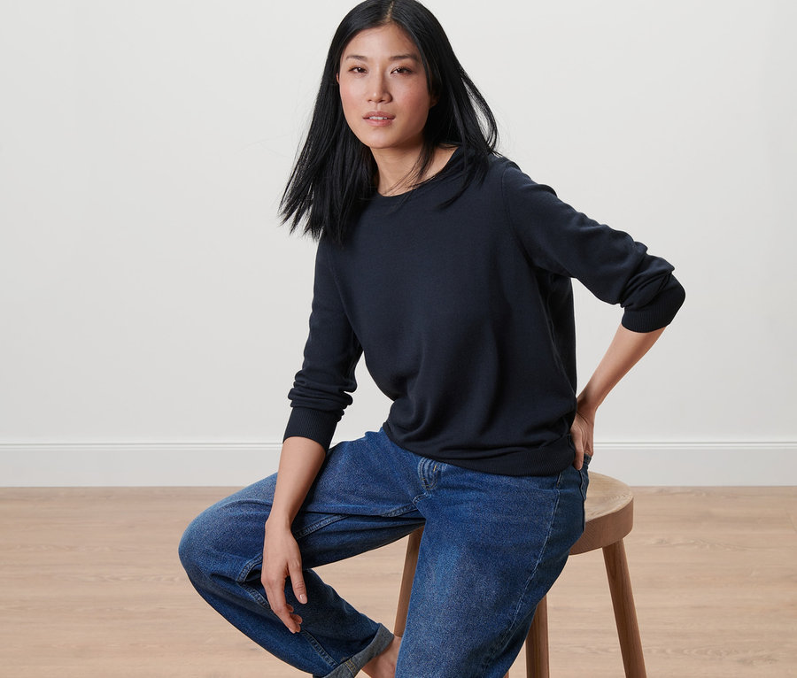 Frau sitzt auf einem Hocker und trägt einen dunkelblauen Merino-Feinstrickpullover.