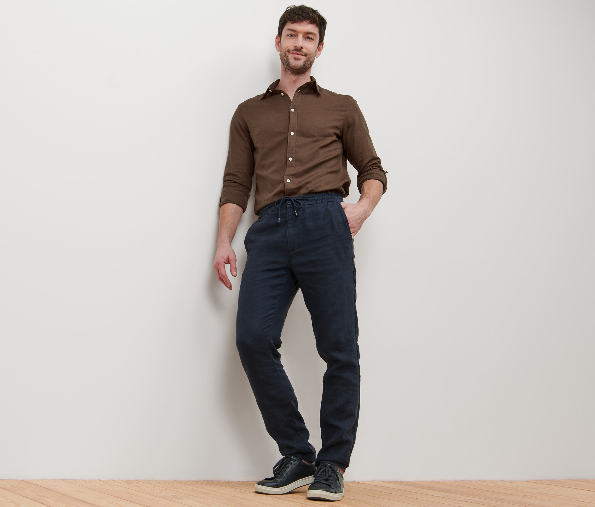 Ein Mann posiert an eine Wand gelehnt in einem braunen Hemd und einer Leinen-Chino.