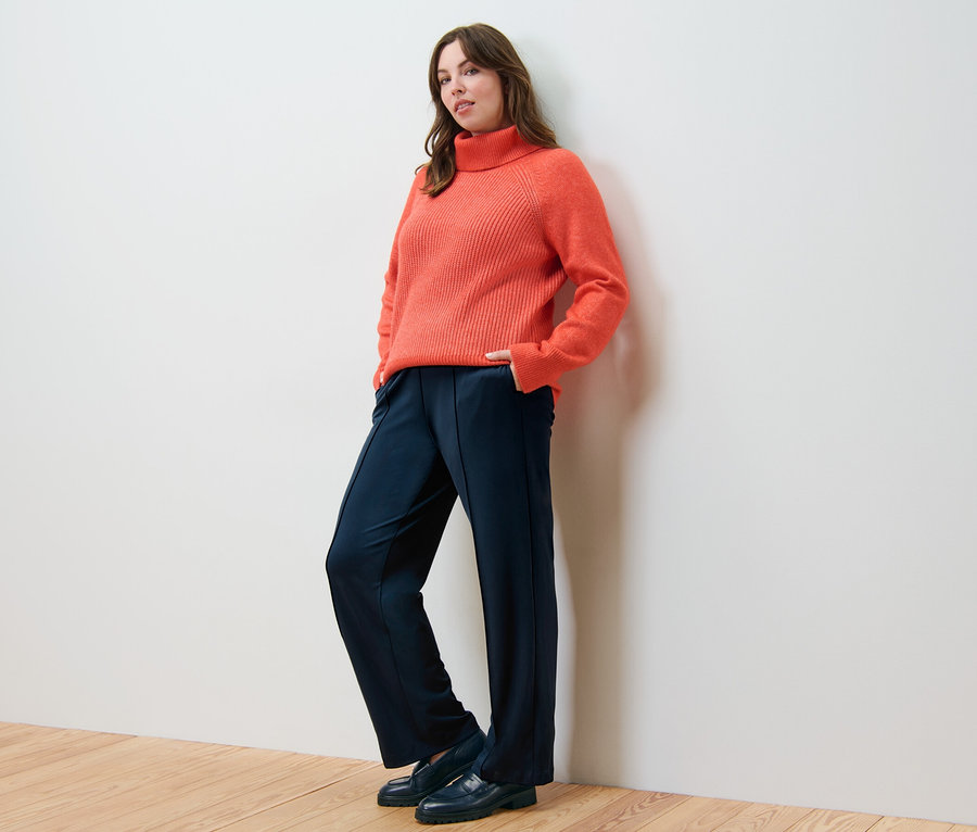 Frau posiert in orangefarbenem Grobstrickpullover mit Rollkragen und Jerseyhose mit Biese.