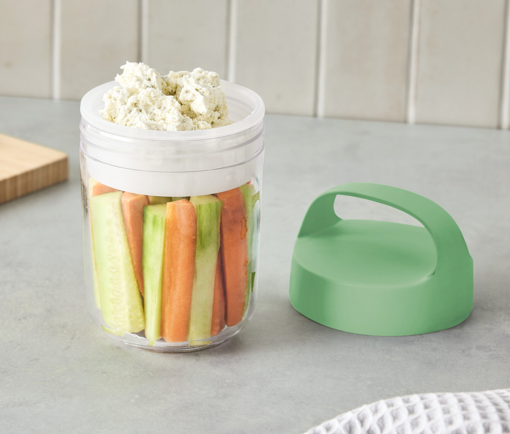 Ein kleiner To-Go-Behälter mit Henkel, gefüllt mit geschnittenem Gemüse und Dip.