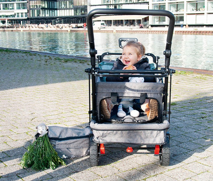 Ein lächelndes Mädchen sitzt in einem grauen Pinolino Klappbollerwagen Add Plus auf einem Gehweg neben einem Fluss.