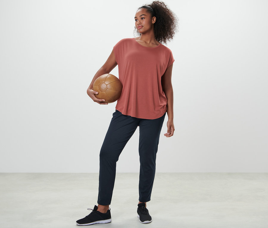 Frau hält einen Fußball, trägt ein terrakottafarbenes Sportshirt und eine Trainingshose.
