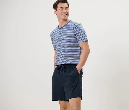 Mann steht und lächelt, er trägt einen blau-dunkelblauen Shorty-Pyjama.