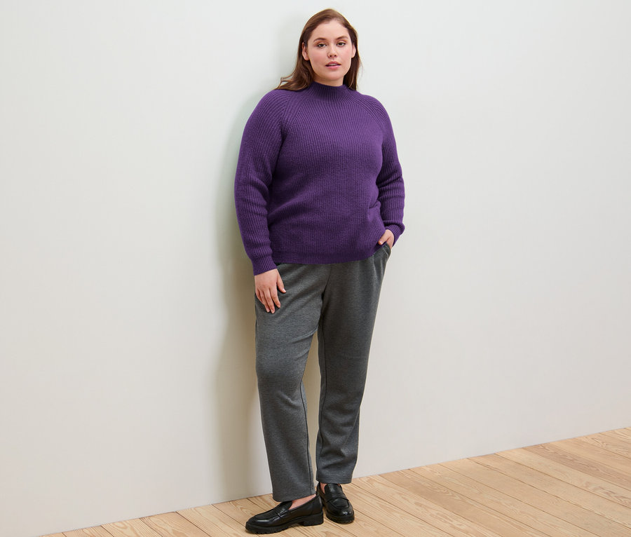 Eine Frau steht mit den Händen in den Taschen ihrer Joggpants und trägt einen lila Grobstrickpullover mit Stehkragen.