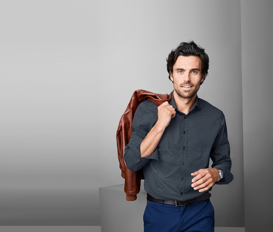 Mann mit brauner Lederjacke über der Schulter posiert in einem Hemd, Modern Fit.