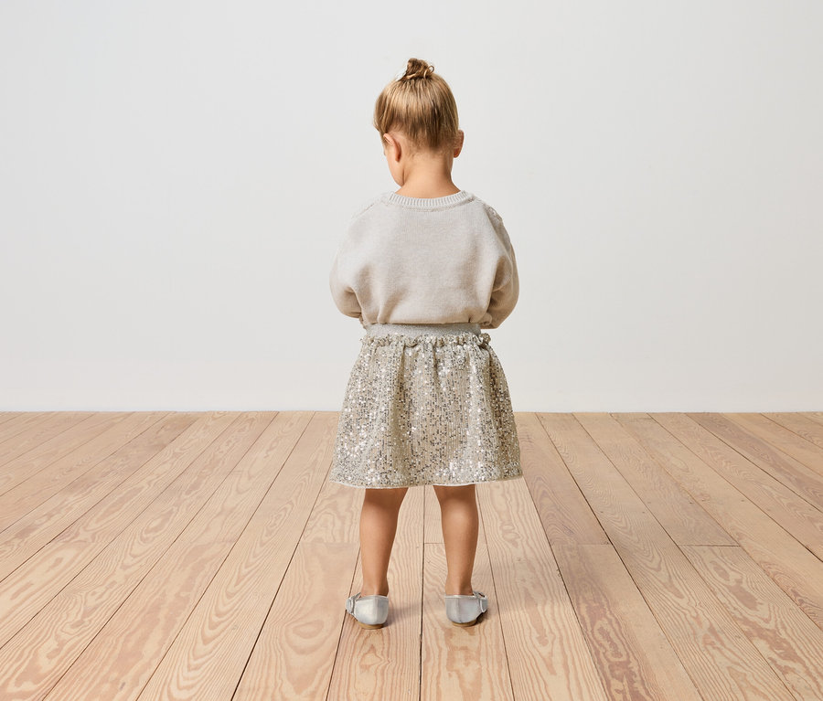 Ein kleines Mädchen steht mit dem Rücken zum Betrachter, es trägt einen glitzernden Rock und einen Pullover.