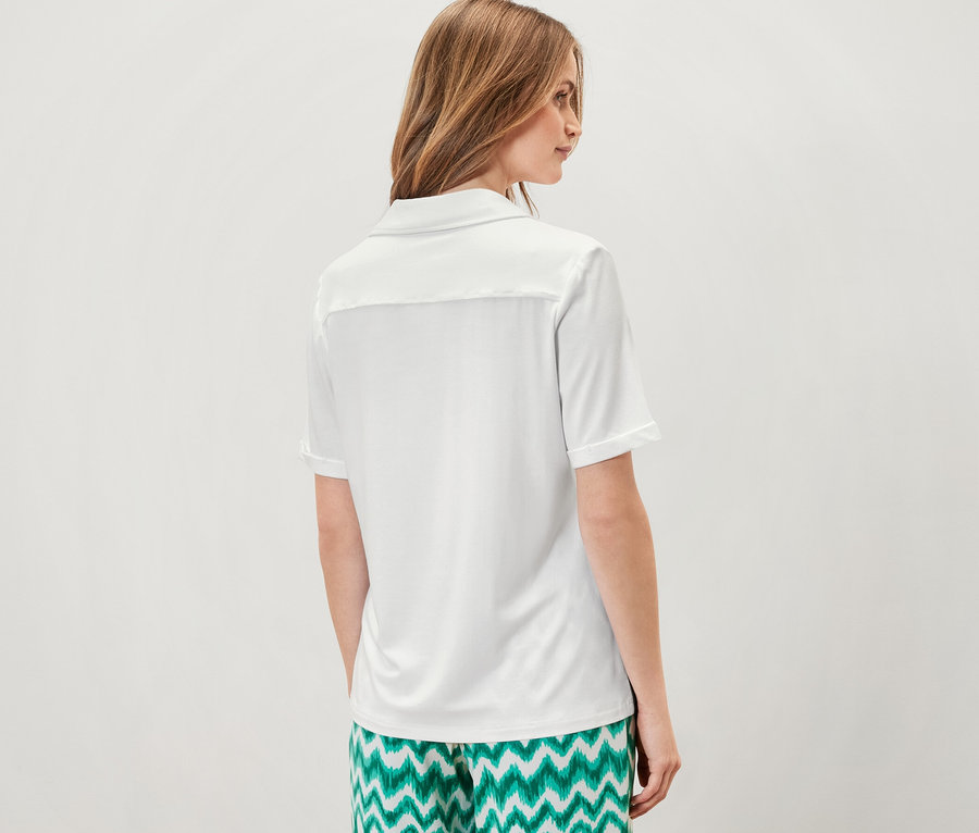 Frau steht mit dem Rücken zur Kamera und trägt ein weißes Blusenshirt mit Knopfleiste und eine grüne, gemusterte Hose.