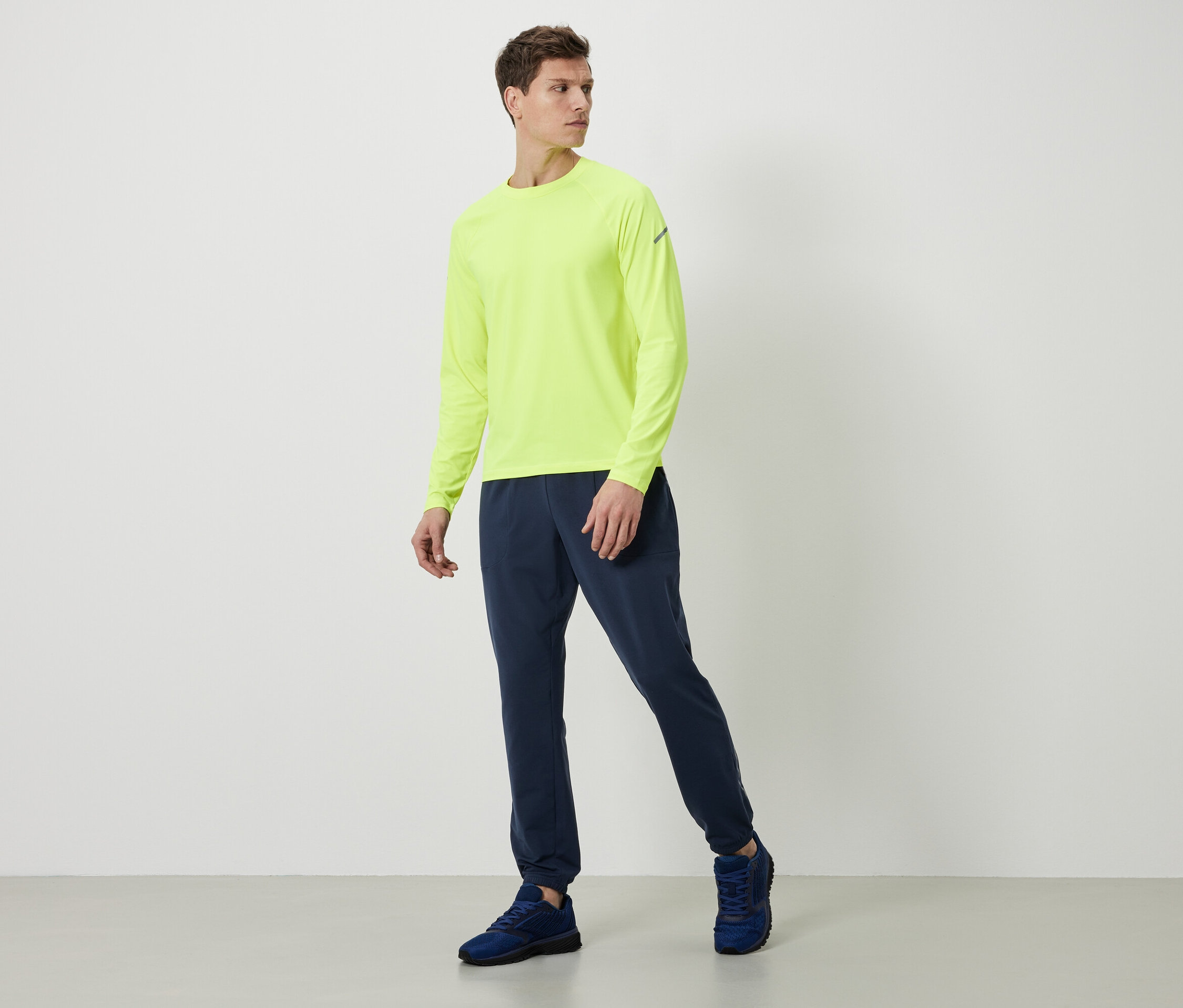 Mann in neon gelbem Langarmshirt, dunkelblauer Trainingshose und blauen Schuhen.