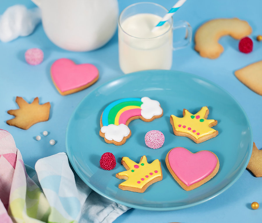 Blauer Teller mit Regenbogen-, Herz- und Kronenkeksen und Bonbons auf hellblauem Hintergrund. Im Hintergrund ein Glas Milch und ein Krug. Chefclub Dessert und Keks-Back-Set.