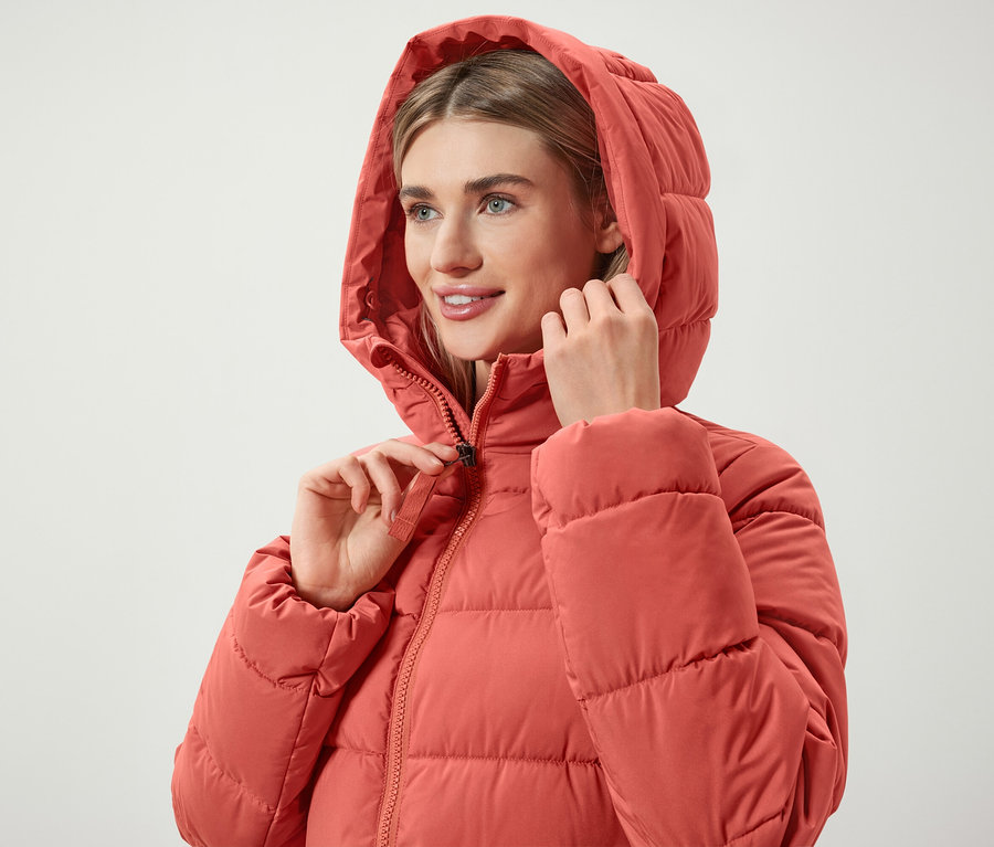 Frau schließt den Reißverschluss einer korallenroten Daunenjacke mit Kapuze.