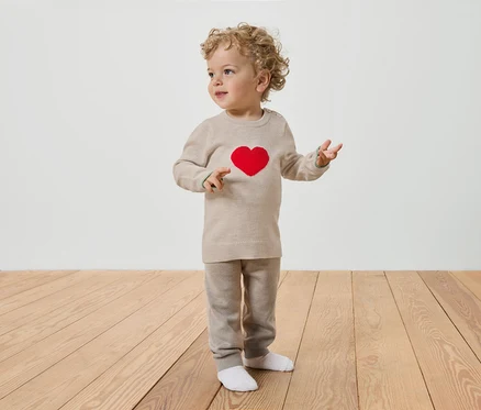 Ein Kleinkind mit lockigem Haar steht auf einem Holzboden und trägt einen beigen Pullover mit einem roten Herz und eine beige Hose.