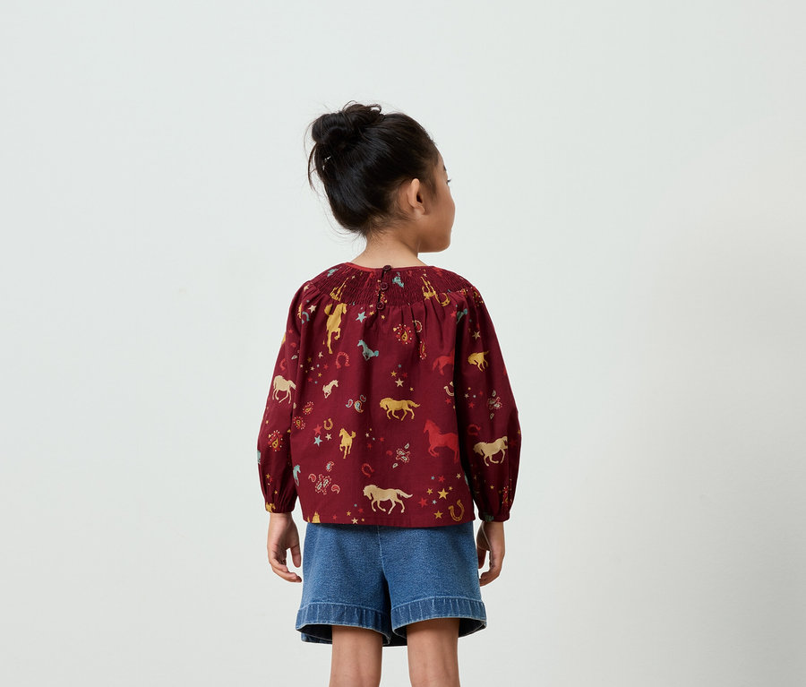 Rückansicht eines Mädchens mit Duttfrisur, bekleidet mit einer weinroten Bluse mit Pferdeaufdruck und blauen Jeansshorts.