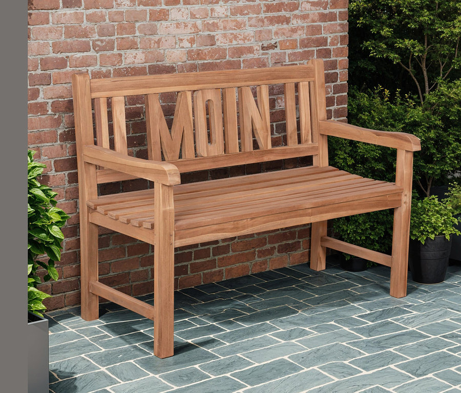 Eine hölzerne Gartenbank mit der Aufschrift "MOIN" steht auf einer gepflasterten Terrasse vor einer Ziegelmauer. Im Titel genannt: deVries Gartenbank »Moin«