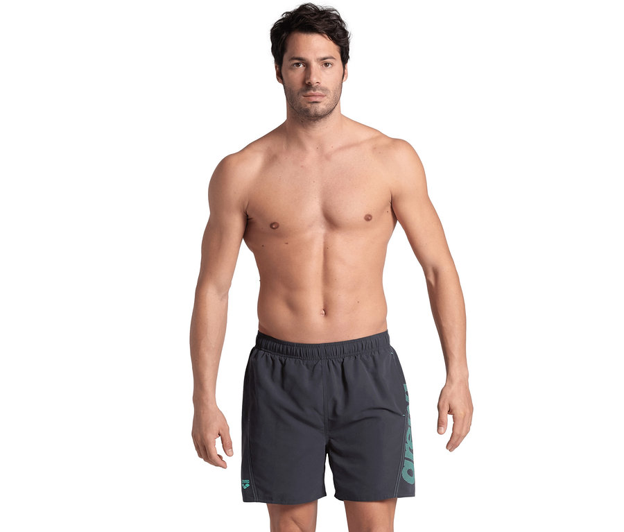 Mann ohne Hemd posiert im Studio und trägt graue Arena Badeshorts Fundamentals Logo.