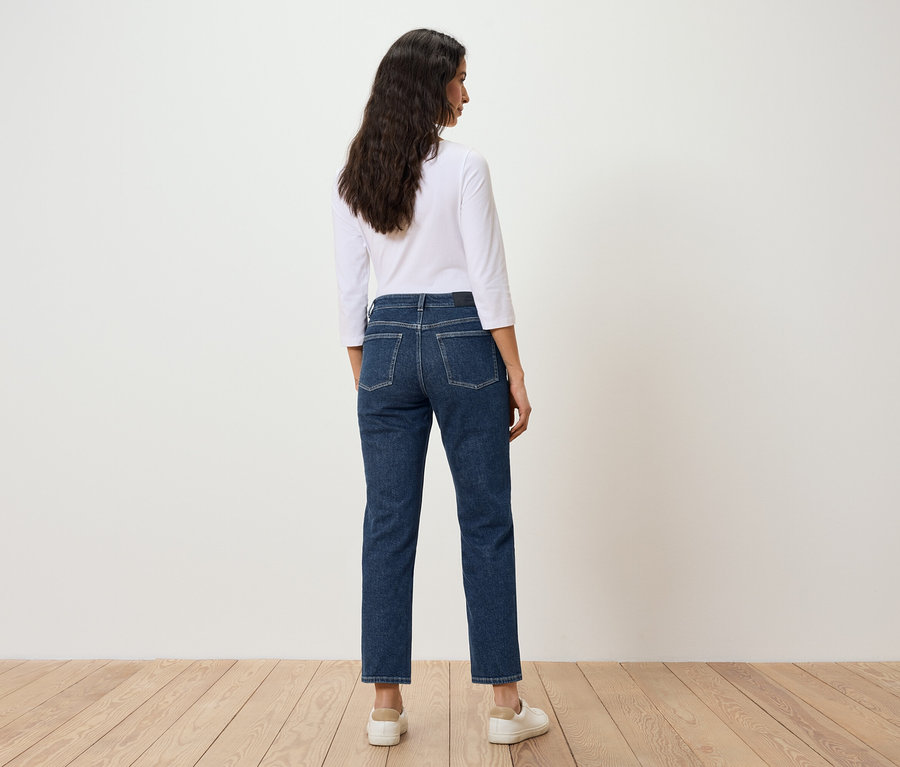 Eine Frau mit dunklen Haaren steht mit dem Rücken zur Kamera und trägt blaue Jeans und ein weißes T-Shirt.