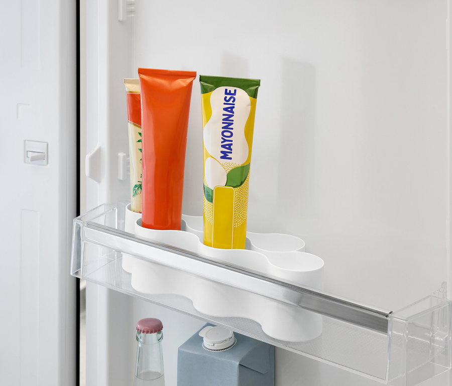 Im Kühlschrank befindet sich ein durchsichtiges Regal mit Tuben mit orangefarbenen, gelben und beigen Etiketten sowie einer Flasche und einem Karton darunter.