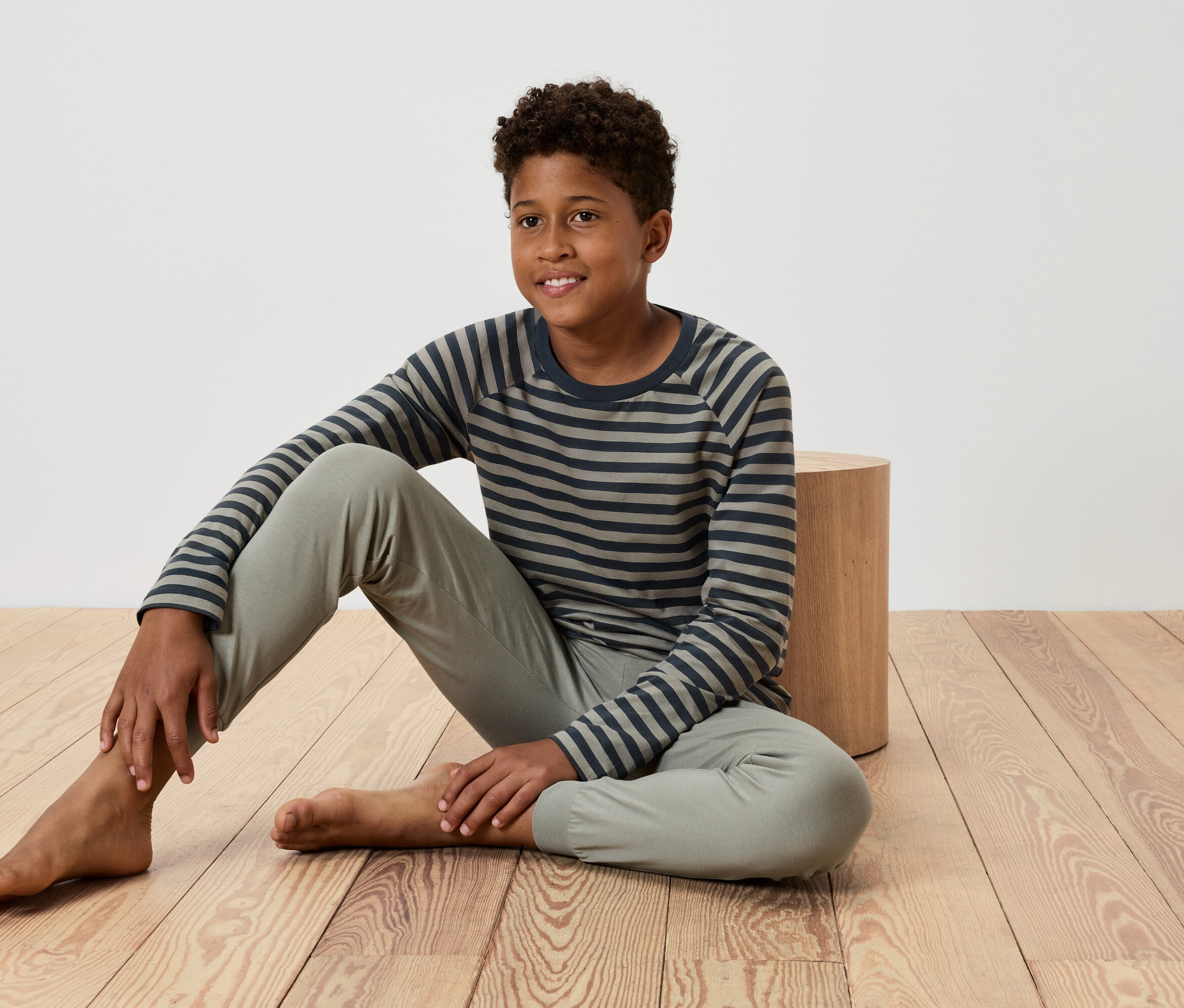 Ein junger Junge sitzt auf einem Holzboden, lehnt sich an einen kleinen Holztisch, trägt ein gestreiftes T-Shirt und eine Hose.