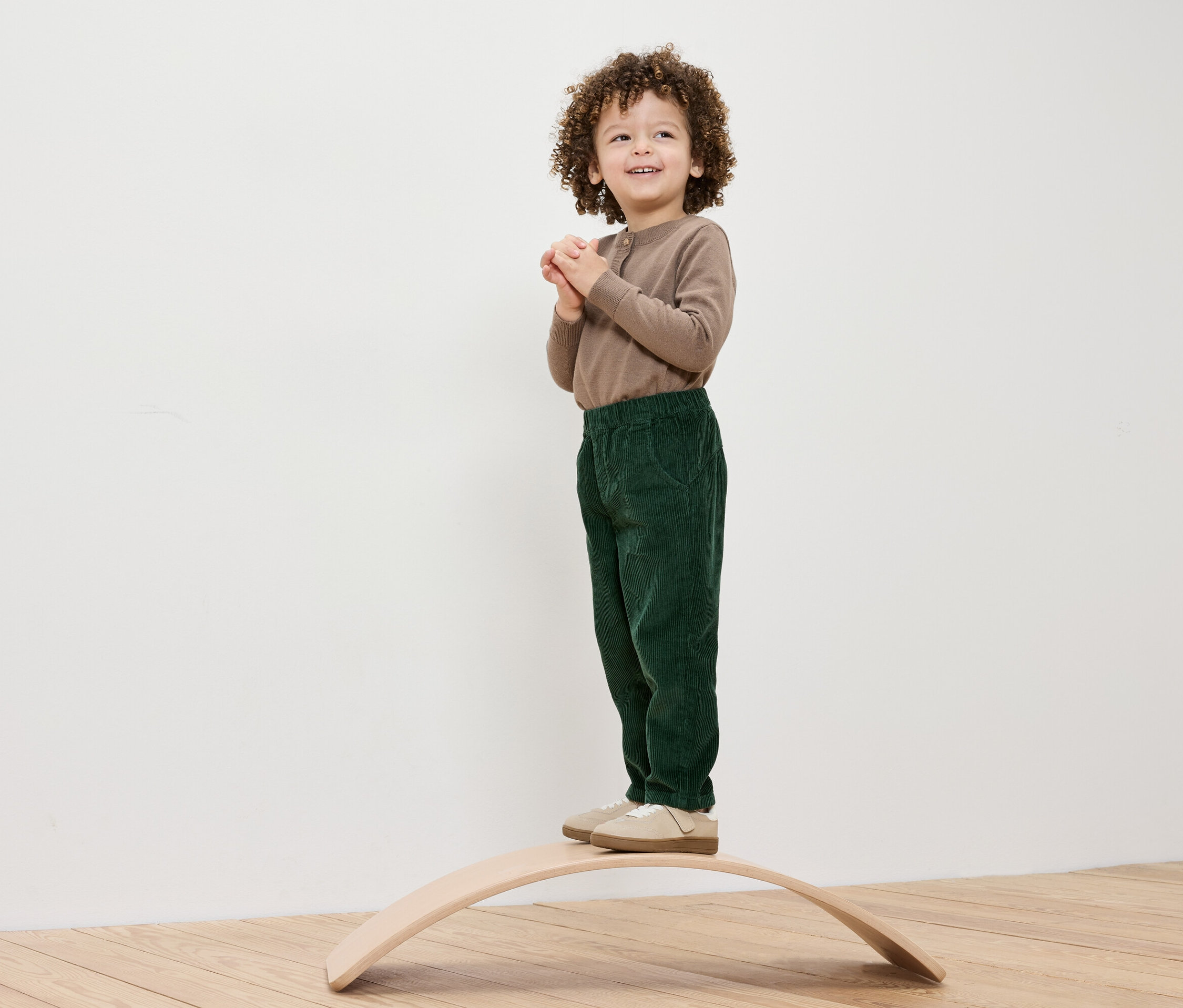 Ein Junge mit lockigem Haar steht auf einem hölzernen Balance Board, er trägt ein braunes Shirt und grüne Hosen.