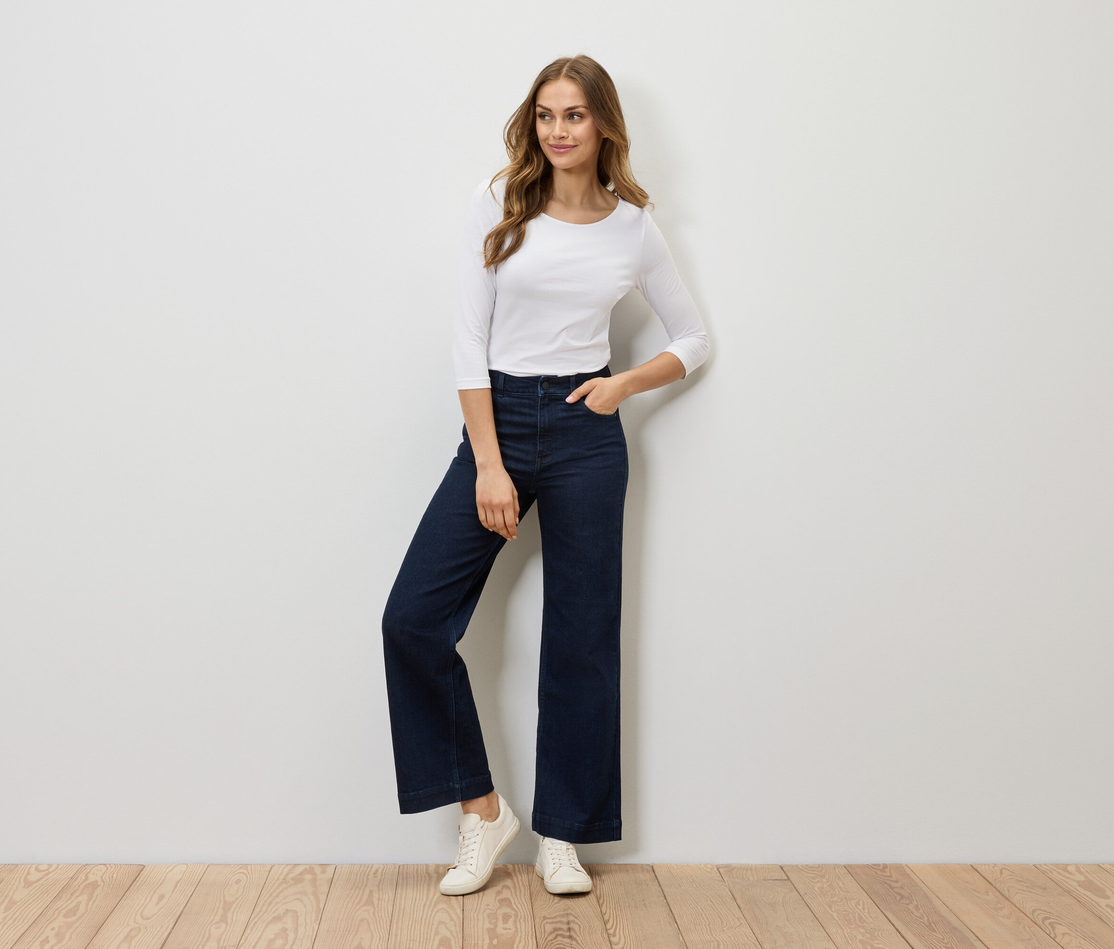 Eine Frau posiert in einem weißen Dreiviertelarm-Shirt, einer dunkelblauen Schlaghose und weißen Turnschuhen.