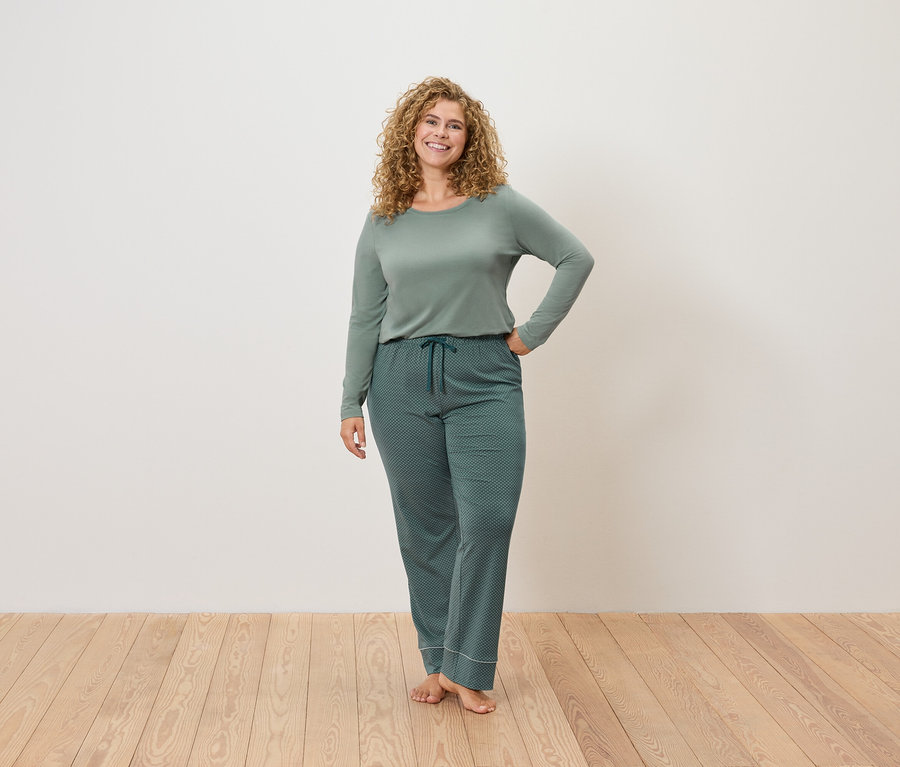 Frau steht und posiert in grüner Pyjamakleidung auf Holzboden vor einer weißen Wand.