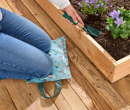 Eine Person kniet auf einem blauen Kniekissen mit Blumenmuster neben einem Hochbeet und benutzt eine kleine Schaufel, um Blumen einzupflanzen.