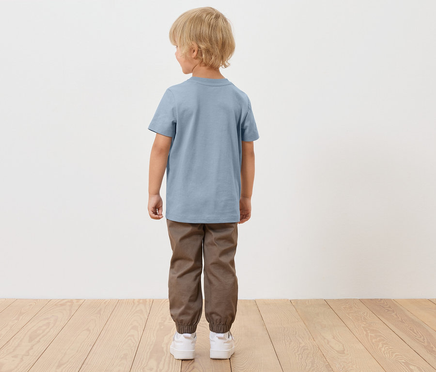 Ein blonder Junge steht mit dem Rücken zum Betrachter und trägt ein blaues T-Shirt, braune Hosen und weiße Schuhe.