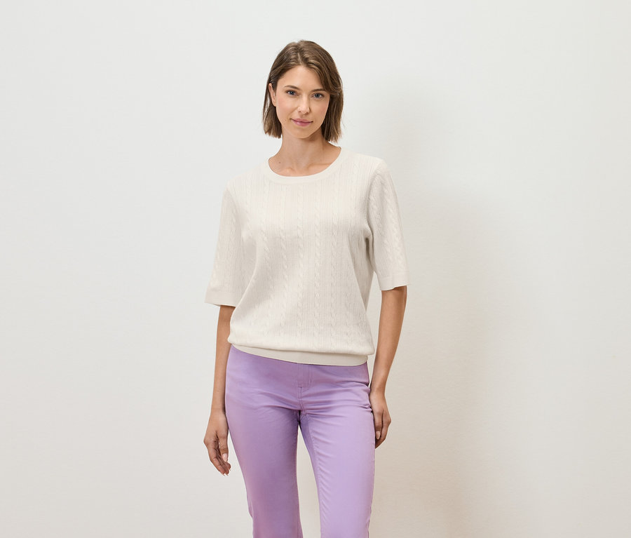 Eine Frau posiert vor einem hellen Hintergrund in einem cremefarbenen Kurzarmpullover und einer lila Hose.