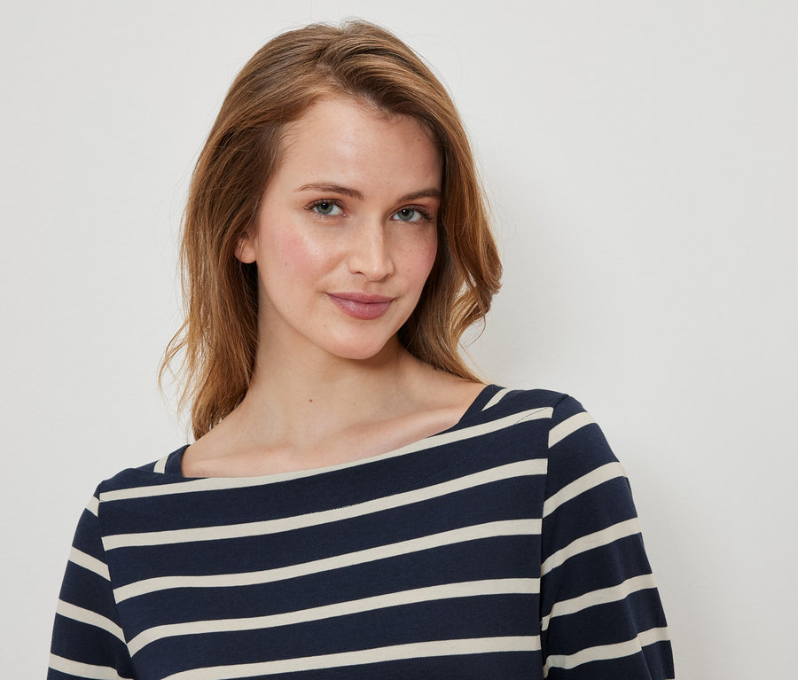 Porträt einer Frau mit braunen Haaren und blauen Augen, die ein blau-weiß gestreiftes Oberteil trägt.