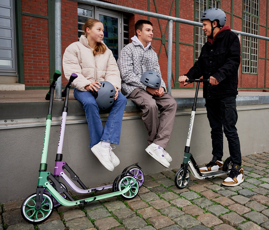 Drei Jugendliche draußen mit HUDORA BigWheel® 205 Rollern. Ein Mädchen sitzt neben einem grünen und lavendelfarbenen Roller, während ein Junge auf einem grünen Roller steht.