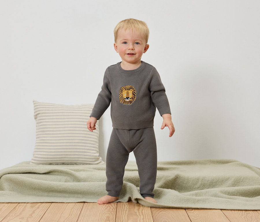 Kleiner Junge steht barfuß auf einer hellgrünen Decke, trägt einen grauen Strickpullover mit Löwenmotiv und eine graue Hose.