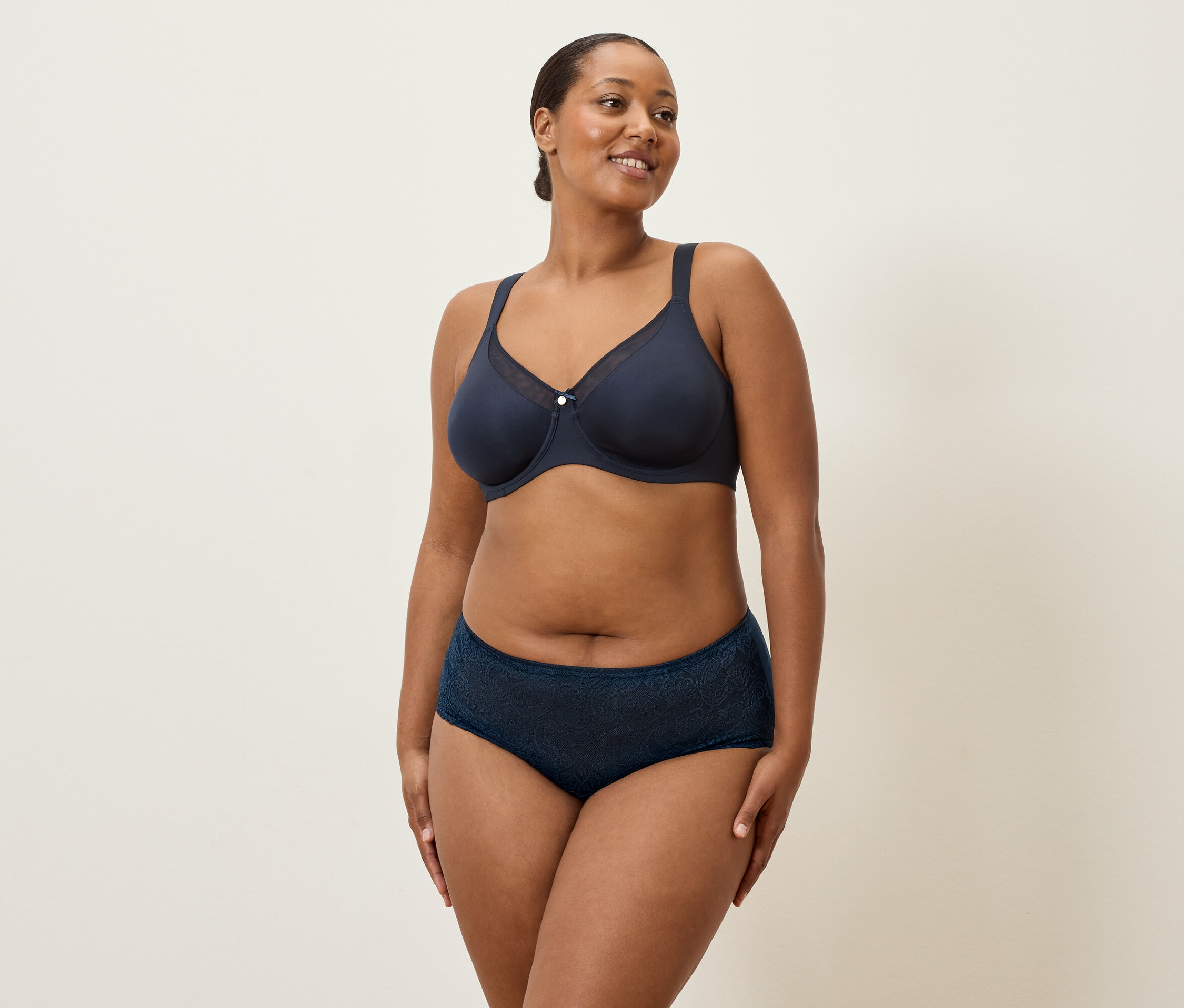 Eine Frau mittleren Alters mit dunklen Haaren und marineblauer Unterwäsche steht vor einem cremefarbenen Hintergrund.