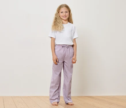 Mädchen mit blonden, lockigen Haaren, weißem T-Shirt und gestreifter, lilafarbener Hose steht barfuß auf einem Holzboden vor einer weißen Wand.