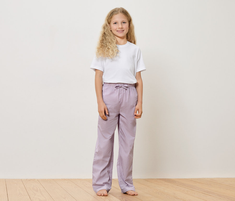 Mädchen mit blonden, lockigen Haaren, weißem T-Shirt und gestreifter, lilafarbener Hose steht barfuß auf einem Holzboden vor einer weißen Wand.